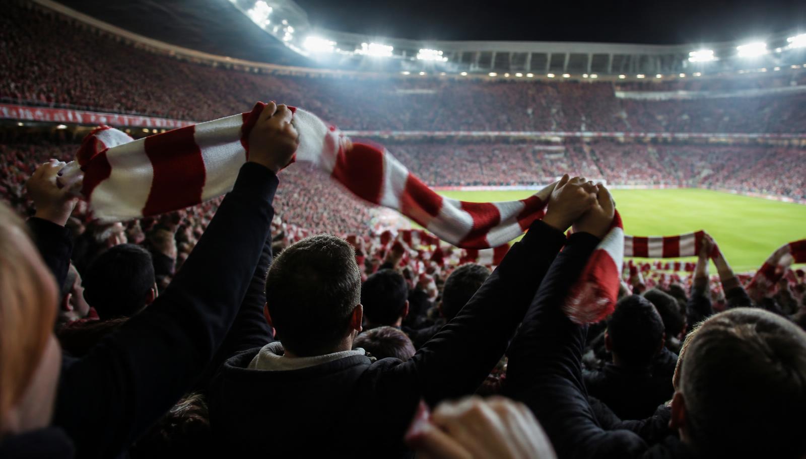 Football supporters holding red and white scarves above their heads in a floodlit stadium